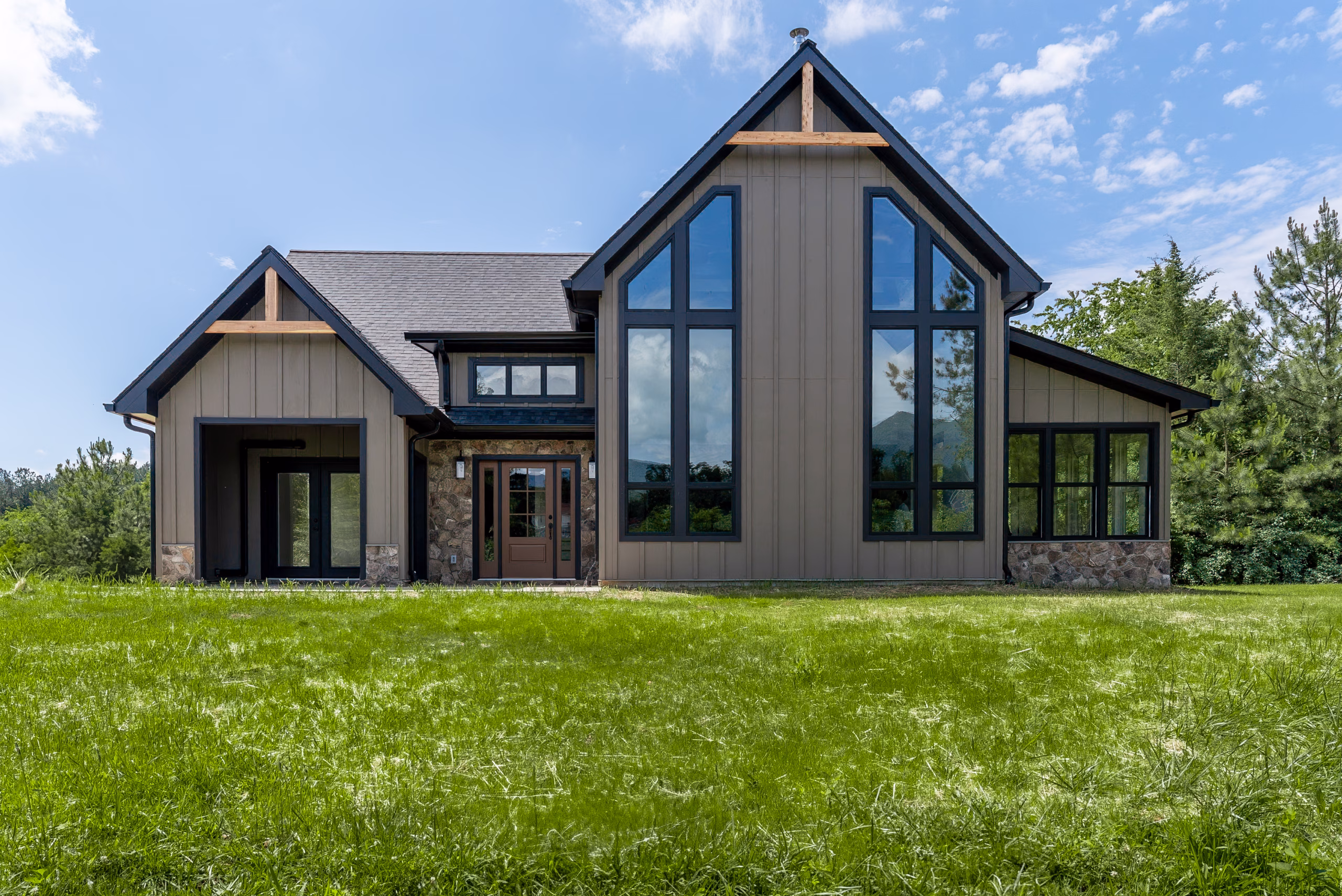 Front exterior view of a beautiful taupe A frame style home with large windows, black trim and gutters. Accents of natural stone surround the door and the lower portion of the foundation and wood accents are in the gables.